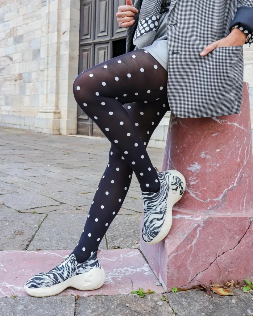Medias negras con lunares blancos, piernas cruzadas frente a una fachada de piedra, look urbano con zapatillas. Black tights with white polka dots, crossed legs in a street style outfit in front of a stone building.