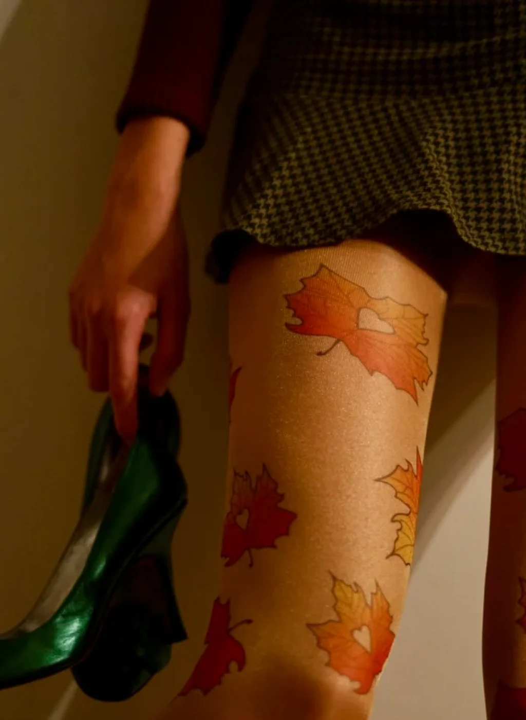 Pantimedias beige con hojas de otoño y falda corta, modelo sosteniendo zapatos verdes | Beige tights with autumn leaves print and short skirt, model holding green shoes