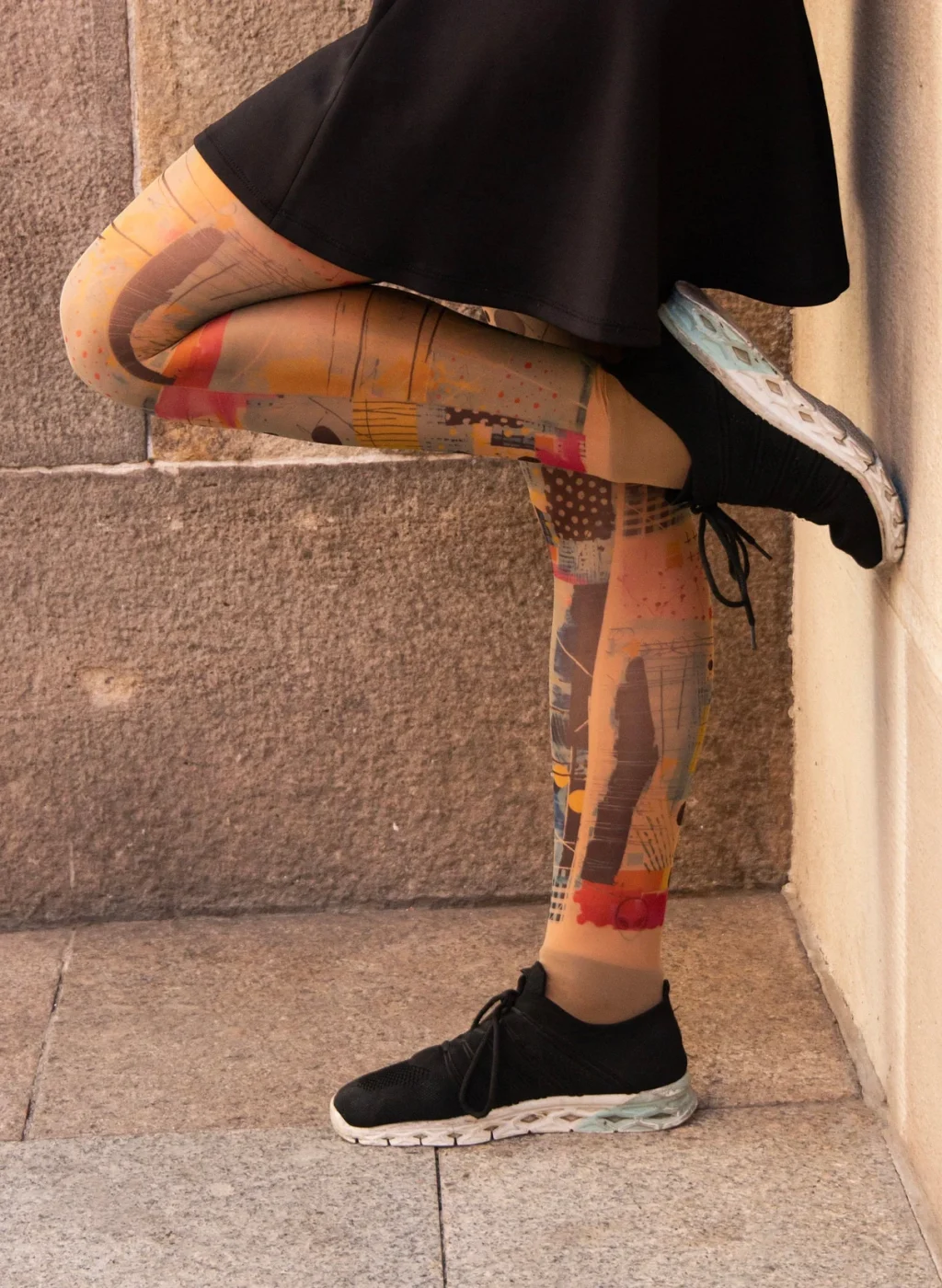 Piernas con pantimedias de arte abstracto apoyadas en pared de piedra con falda negra | Legs wearing abstract art tights leaning against stone wall with black skirt