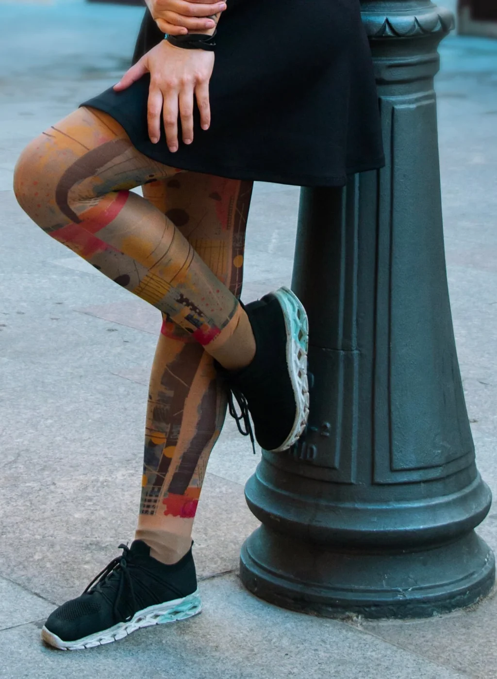 Piernas con pantimedias de arte abstracto y colores vivos, sentada junto a farola urbana | Legs in vibrant abstract art tights sitting beside street lamp