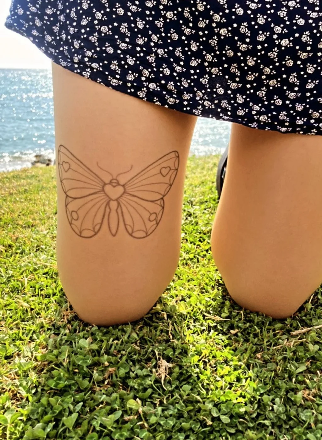 Sheer nude tattoo tights with large butterfly design on the thigh, outdoor grass background | Pantimedias nude semitransparentes con diseño de mariposa grande en el muslo, fondo de césped exterior