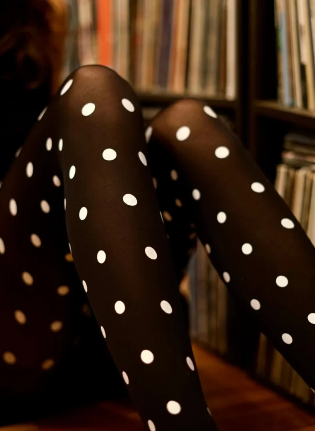 Medias negras con lunares blancos, piernas cruzadas junto a una mujer sentada en un ambiente cálido con estantería de discos de vinilo. Black polka dot tights, crossed legs next to a seated woman in a warm cozy setting with a vinyl record shelf.