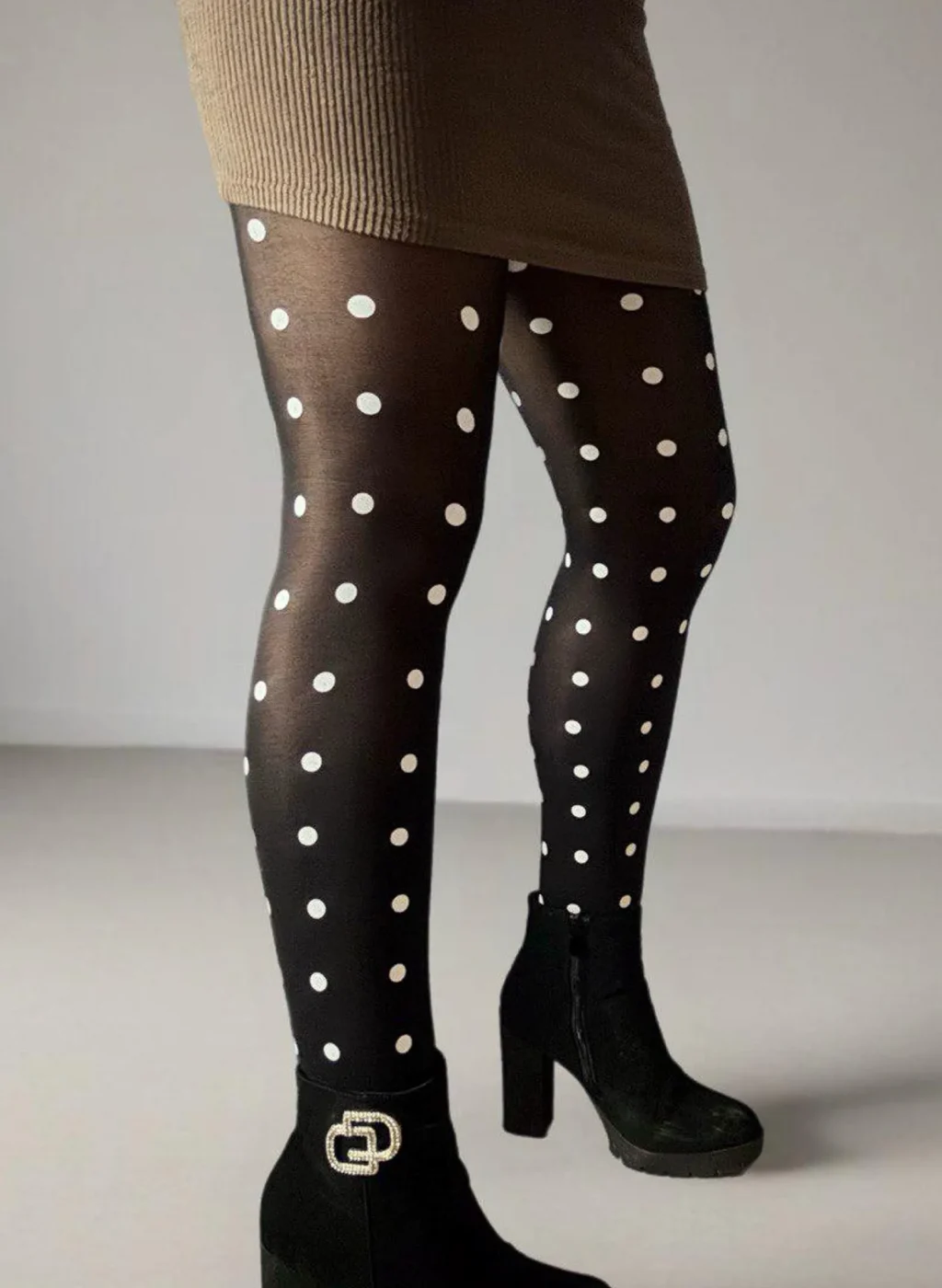 Medias negras con lunares blancos, piernas de pie con vestido ceñido beige y botines negros sobre fondo neutro. Black polka dot tights, standing legs with fitted beige dress and black ankle boots on a neutral background.