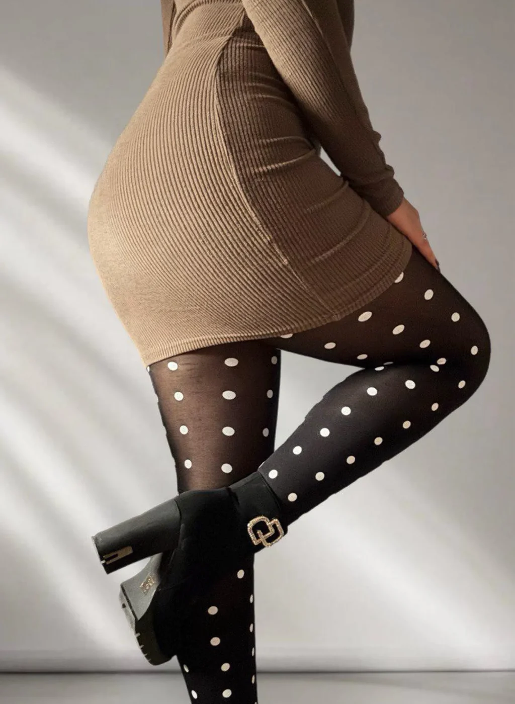 Medias negras con lunares blancos, piernas dobladas con vestido ceñido beige y botines negros, iluminadas por luz natural lateral. Black polka dot tights, bent legs in a fitted beige dress and black ankle boots, lit by soft side natural light.