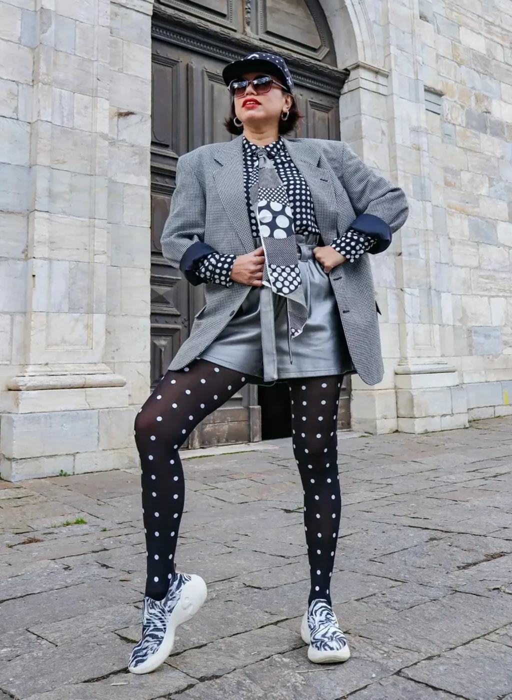 Medias negras con lunares blancos en look urbano con blazer gris, gafas de sol y zapatillas, posando frente a una fachada de piedra. Black polka dot tights in a street style outfit with grey blazer, sunglasses and sneakers, posing in front of a stone building.