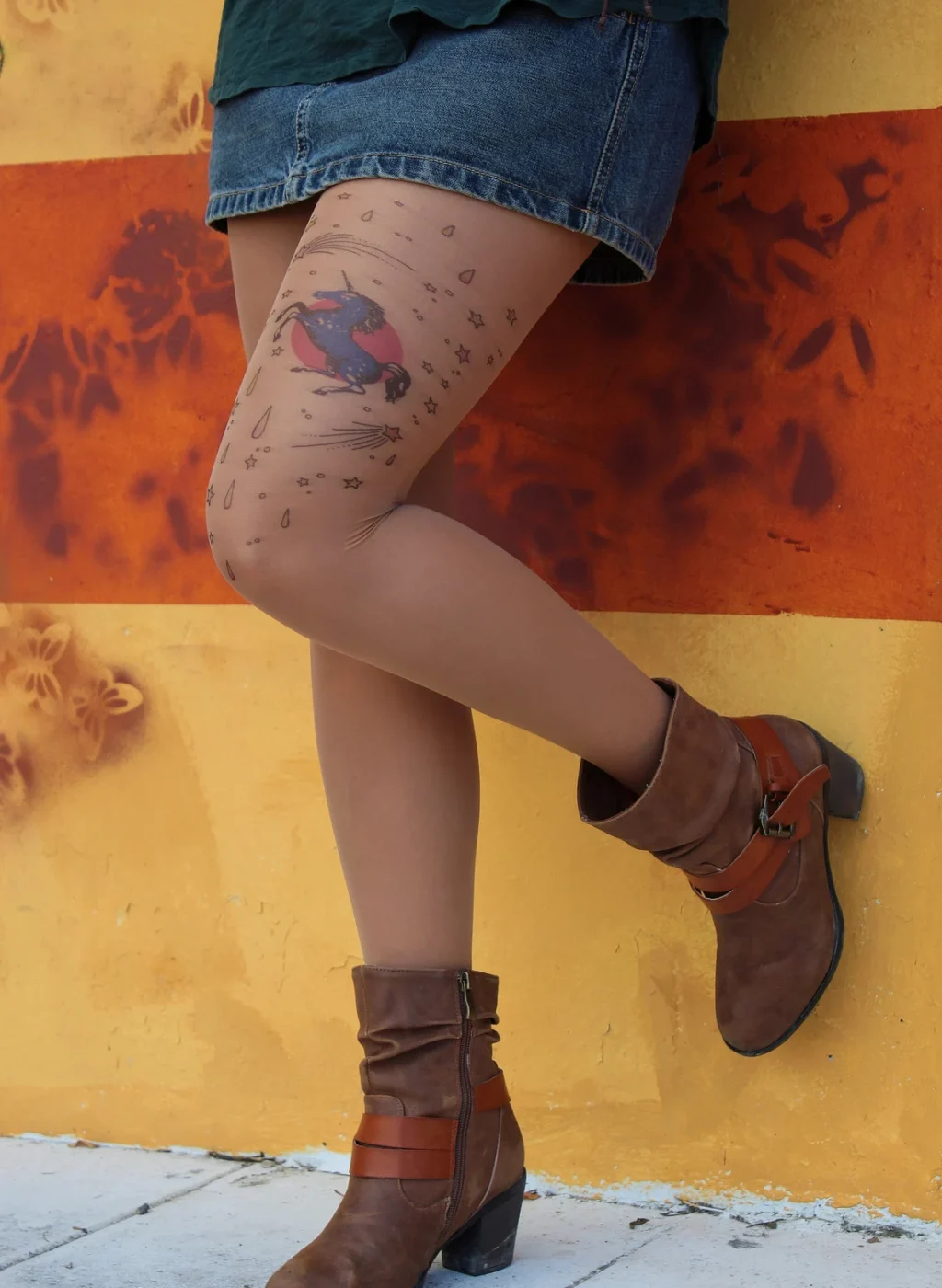 Leg wearing sheer pantyhose with a colorful unicorn tattoo design, outdoors against an orange wall | Pierna con pantimedias transparentes con diseño de unicornio tatuado, en exterior junto a una pared naranja