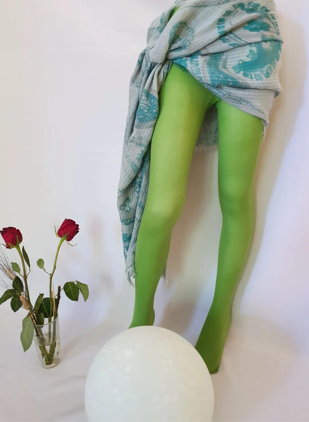 Pantimedias verde manzana en piernas posando junto a flores rojas y esfera decorativa sobre fondo blanco | Apple green tights with legs posing beside red flowers and decorative sphere